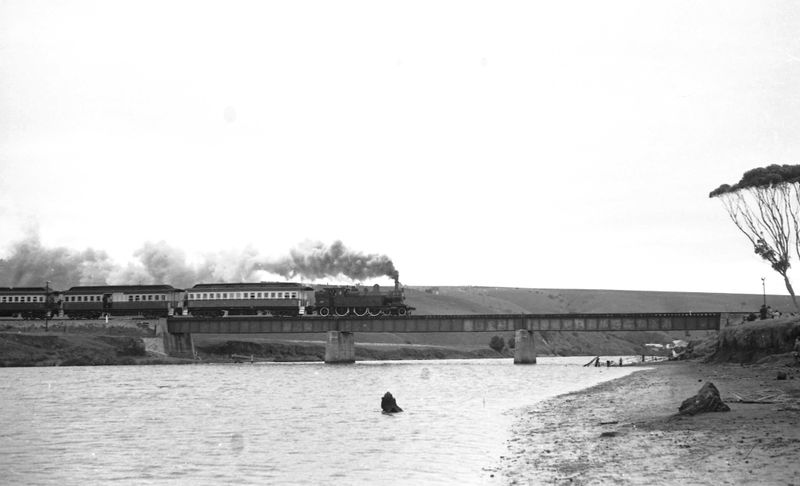 Black-and-white photograph of a steam locomotive hauling passenger carriages across the Noarlunga rail bridge over the Onkaparinga River, seen from the sandy riverbank