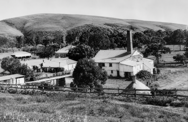 Black-and-white photograph looking down on the Horseshoe Mill at Noarlunga — a cluster of white rendered buildings with a tall chimney stack, set in a creek-side valley beneath a rounded hill
