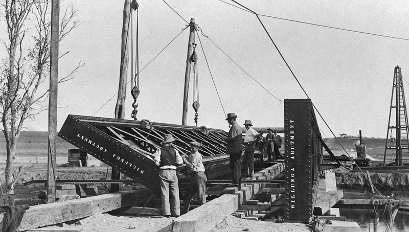 Black-and-white photograph of workmen constructing the Noarlunga railway bridge, with a crane and girder section visible and workers standing on the structure