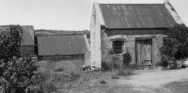 Black-and-white photograph of stone farm outbuildings at Tapley's Hill — a rubble-stone cottage with corrugated iron roof and timber door, ruins of another structure to the left, hills behind