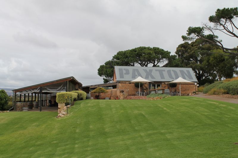 Colour photograph of a stone building with a corrugated iron roof bearing the word PAXTON in large block letters, with two white market umbrellas outside and timber wine barrels at the left side. A modern open pergola structure extends to the left, and a mown lawn fills the foreground beneath an overcast sky.