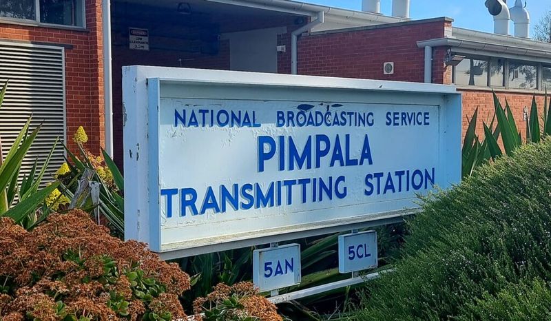 Colour photograph of the station entrance sign reading National Broadcasting Service — Pimpala Transmitting Station — 5AN — 5CL, mounted in front of a red brick building