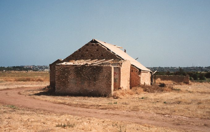 Colour photograph of a roofless stone and brick ruin standing in a bare dry paddock, walls partially collapsed, with suburban development visible in the distant background