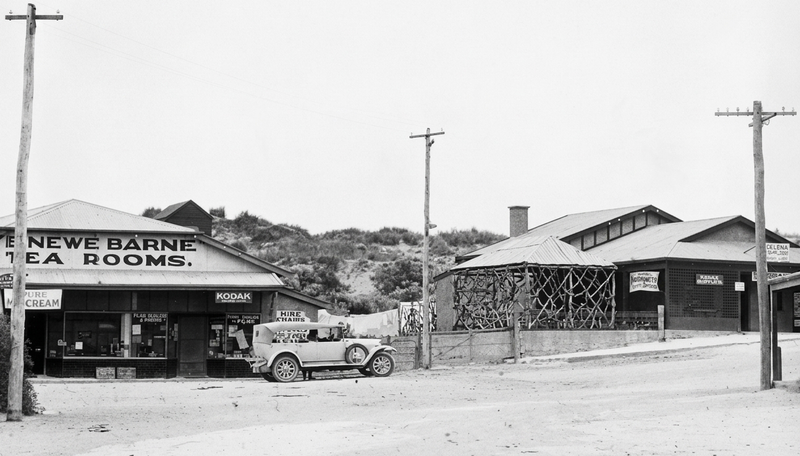 Black-and-white photograph of a dusty Port Noarlunga street in the 1920s — a shop with tea rooms signage on the left and a timber-frame goods shed on the right, a car on the dirt road