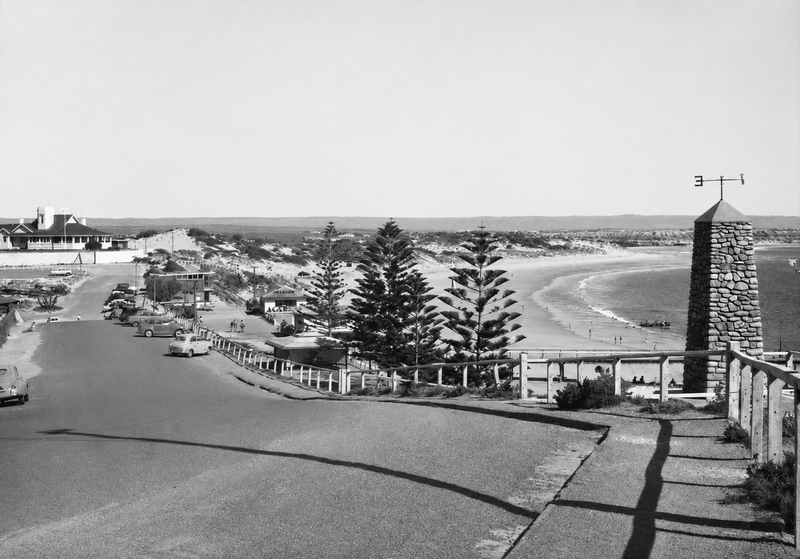 Black-and-white photograph looking down over the Port Noarlunga foreshore — The Esplanade with parked cars and Norfolk Island pines, the beach curving away, and a stone cairn with weathervane in the right foreground