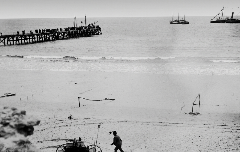 Black-and-white photograph of Port Noarlunga beach from above, showing people crowded on the jetty extending into the surf, vessels at anchor offshore, and a figure on the beach in the foreground