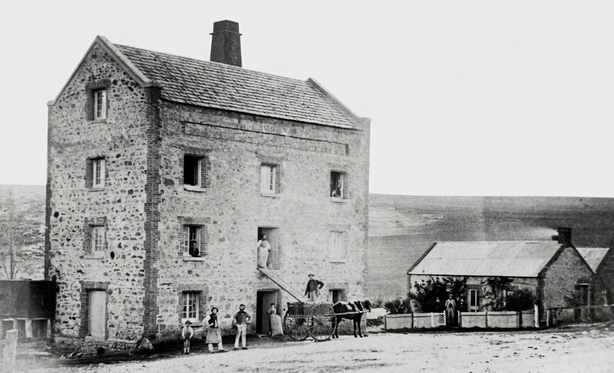 Black-and-white photograph of a tall four-storey stone flour mill building with a chimney, a horse and cart at the base, workers visible at the entrance, and a smaller stone building to the right