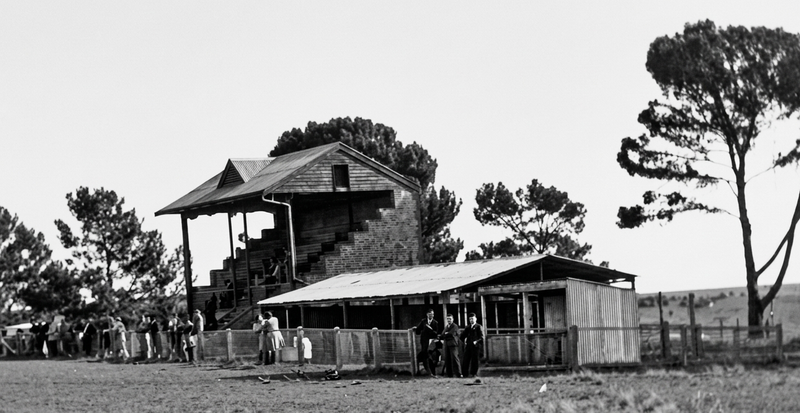 Black-and-white photograph of Reynella Oval's grandstand and changing rooms — a two-storey timber grandstand with external staircase and a lower dressing shed, pines to the right