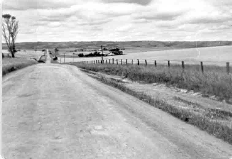 Black-and-white photograph of an unsealed dirt road running away from the camera over a low rise, a post-and-wire fence along the right-hand edge and open rolling farmland under a cloudy sky beyond