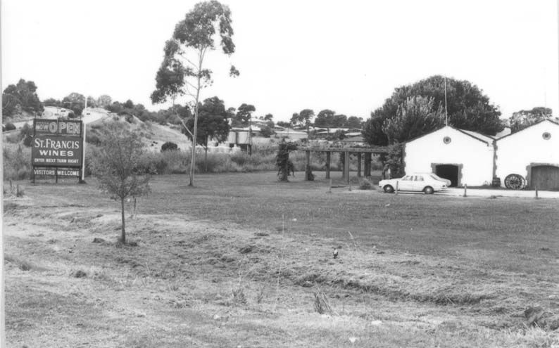 Black-and-white photograph of St Francis Winery — a white-rendered cellar building with a large roadside sign reading Now Open — St Francis Wines — Visitors Welcome, a car parked beside it, the Reynella hills behind