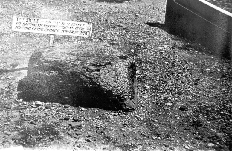 Black-and-white photograph of a dark rocky outcrop embedded in bare earth beside a small placard reading that the stone was used as an anchor for hauling stones by bullocks in the building of the church tower in 1850