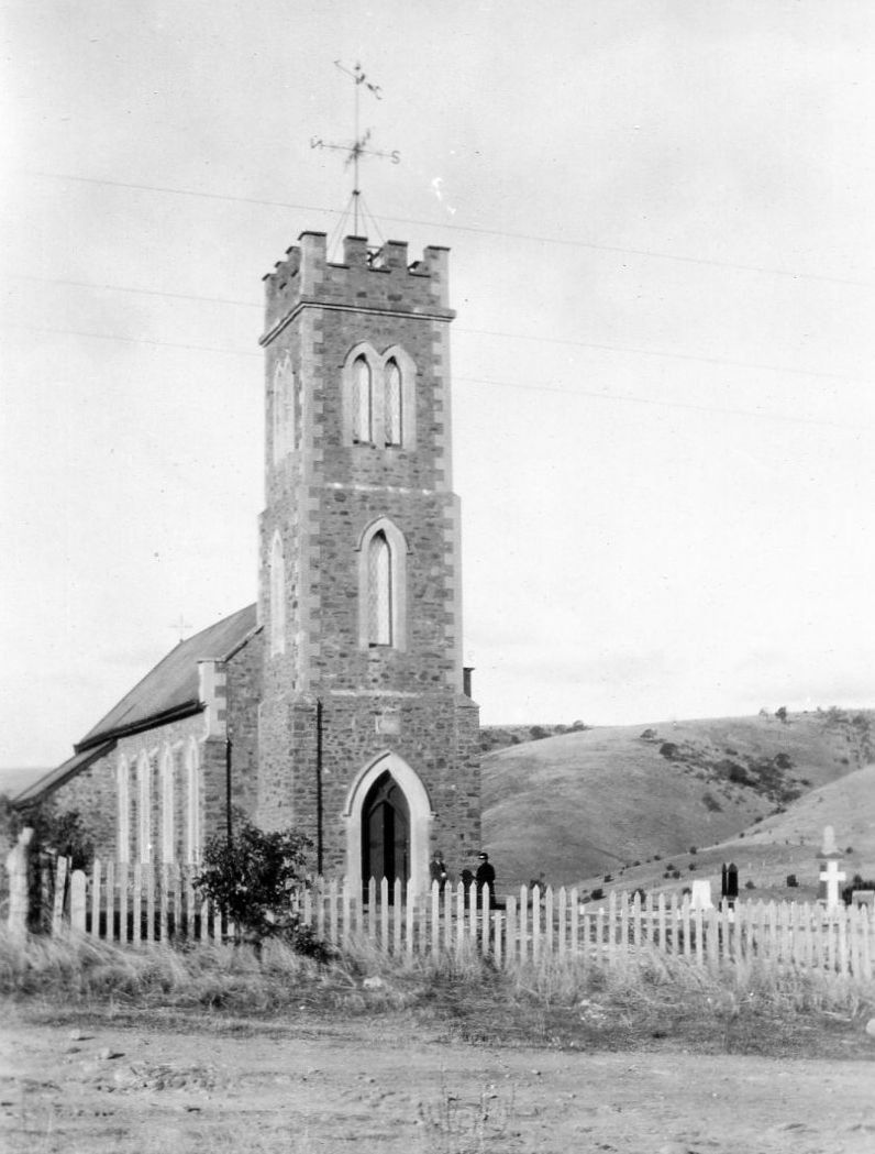 Black-and-white photograph of a stone church with a tall castellated tower topped by a weather vane, a pointed arched doorway and Gothic windows, a white picket fence in the foreground and rolling hills behind