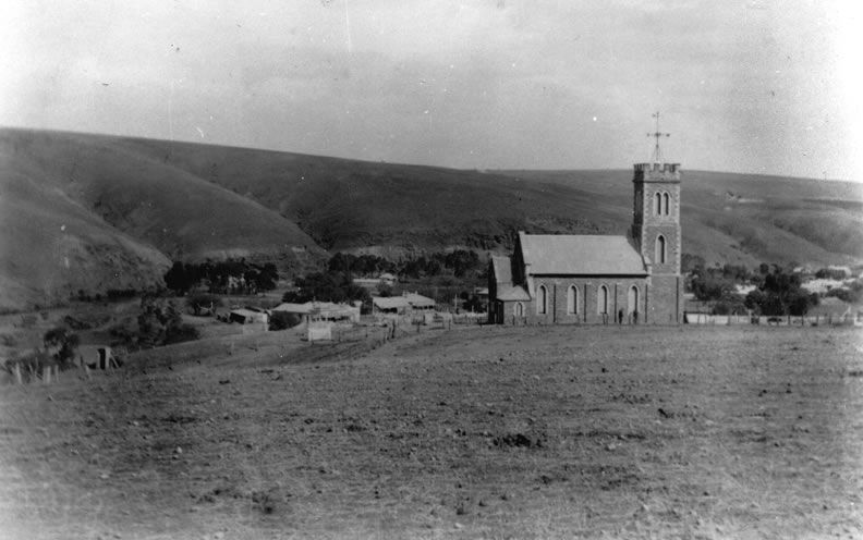 Black-and-white photograph of St Philip and St James Church on a bare hilltop with the Onkaparinga River valley and the scattered buildings of Noarlunga township visible below and rolling hills beyond