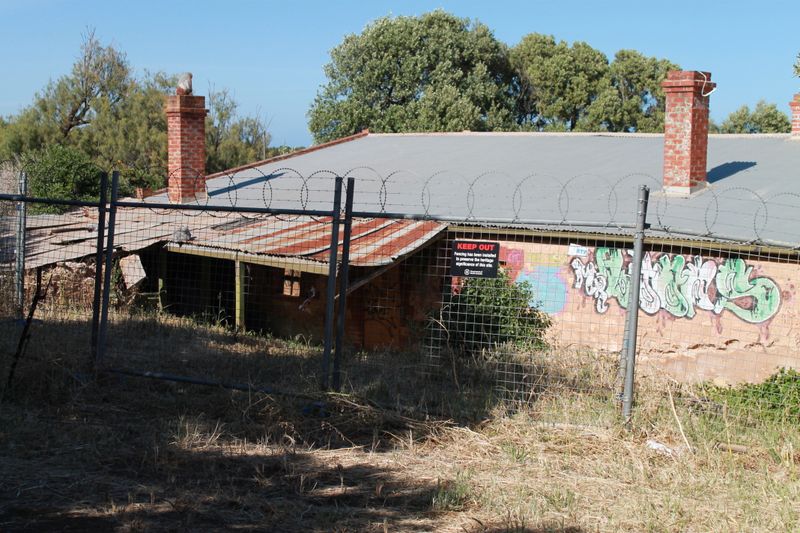 Colour photograph of a long single-storey limestone farmhouse with two tall brick chimneys, a hipped iron roof and a sagging verandah, fenced off behind wire topped with barbed wire, graffiti and a 
