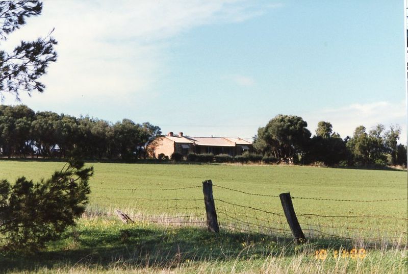 Colour photograph of a low stone farmhouse with a red roof sitting among trees on a low rise, seen across a green paddock with a sagging barbed-wire fence and weathered timber posts in the foreground under a pale blue sky