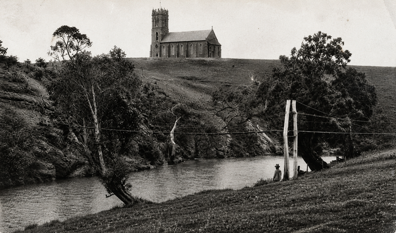 Sepia photograph of the Onkaparinga River with a person sitting on the bank and the old wire-rope crossing mechanism visible; St Philip and St James Church stands prominently on the hilltop above