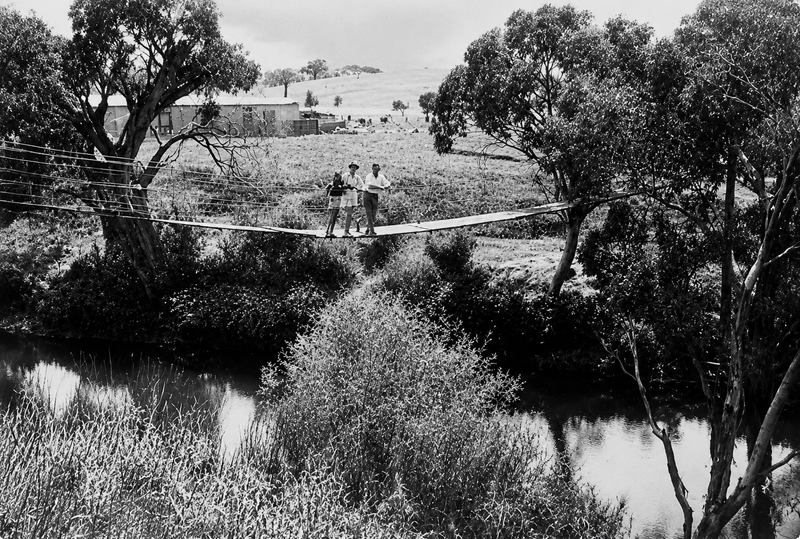 Black-and-white photograph of two people standing on the wire-rope swing bridge suspended over the Onkaparinga River, with gum trees on both banks and a farmhouse in the paddock beyond