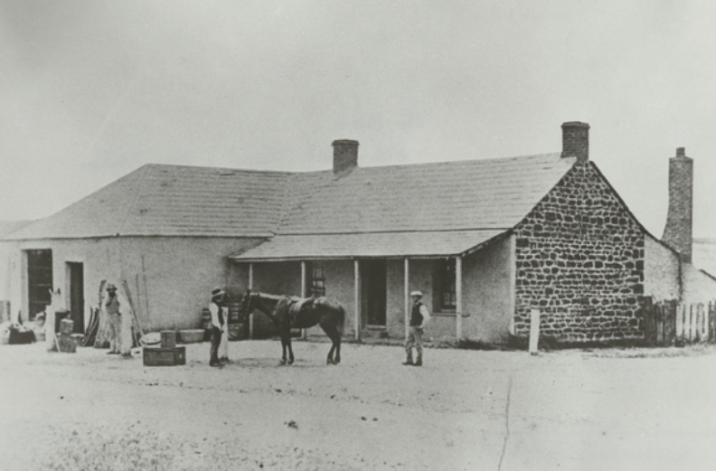Single-storey stone and rendered building with a hipped roof, a verandah and two chimneys; a saddled horse and several figures stand on the dirt road in front, with crates and goods stacked at the left and a paling fence at the right
