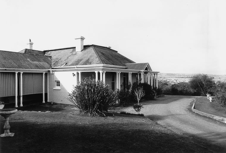 Black-and-white photograph of a single-storey house with a hipped slate roof and two chimneys, a columned verandah across the front and a lower iron-roofed wing to the left, a curving gravel drive and garden with a stone urn in front, and open plain visible in the distance to the right
