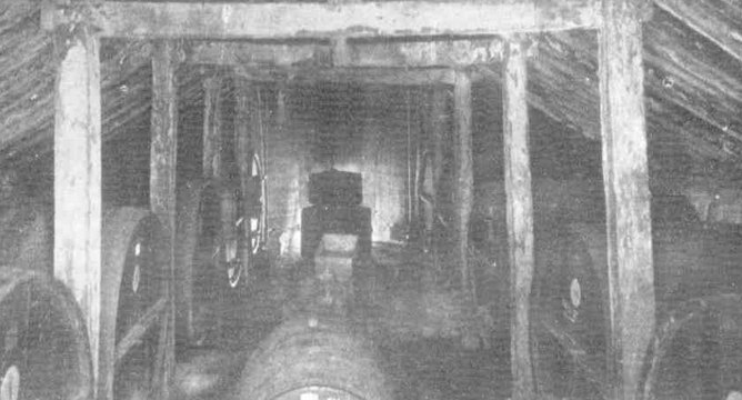 Black-and-white interior photograph looking along the length of the Old Cave cellar, timber roof beams forming an apex overhead and large wine barrels stacked along both sides fading into darkness