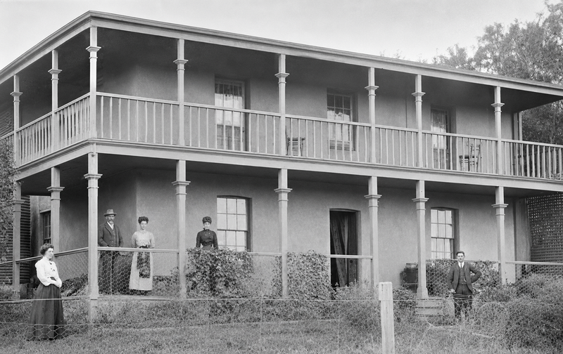 Black-and-white photograph of a two-storey rendered homestead with wide verandahs on both levels, four people standing on the ground floor verandah and lawn in front — The Pines, Happy Valley, 1907