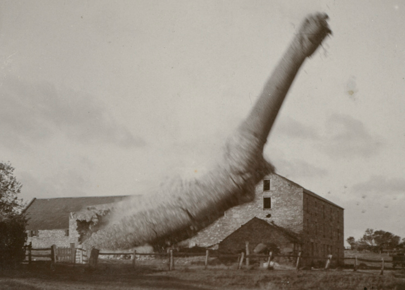 Sepia photograph capturing the demolition of White's Mill chimney — a tall brick chimney caught mid-fall, toppling to one side with a billowing cloud of dust and debris, the stone mill building visible to the right in the background