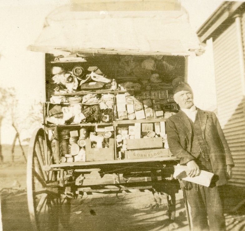 Sepia photograph of a man in a dark three-piece suit and a cap standing at the open side of a horse-drawn van, an order book in his hands; the van