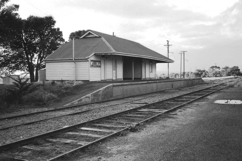 Black-and-white photograph of Willunga station building — a long single-storey weatherboard structure with a wide cantilevered roof over the platform, Willunga nameboard visible, track in the foreground